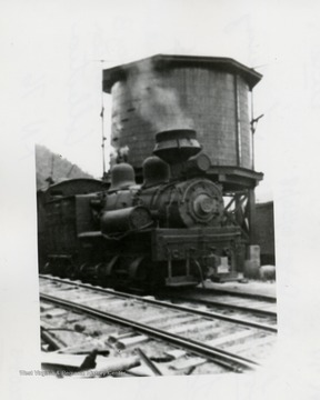 Shay train engine in front of a water tank.  