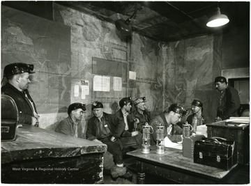 Men sitting down on benches with mine diagrams on the walls. Joe Akers is on the right.