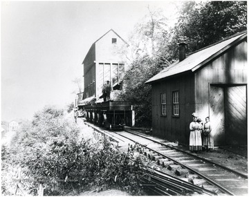 Coal cars with two men standing on top. Lady with two small children are standing, waiting next to a building. 