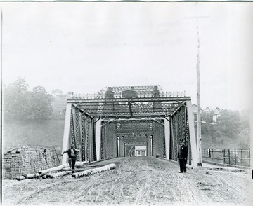 View of the bridge spanning the Monongahela River. Two men standing in front of bridge. One man wearing a suit and a badge. To the right of bridge is a pedestrian walkway. 