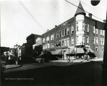View of High Street from the corner of Pleasant Street and High Street in Morgantown, W. Va. Automobiles can be seen parked along side the street, and a person riding a bicycle near the corner. 