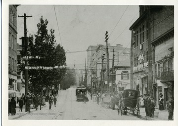 Postcard of High Street in Morgantown, West Virginia. People are walking on the sidewalks of High Street while a trolley is heading toward South High Street and a horse-drane carriage is parked near an unidentified store.