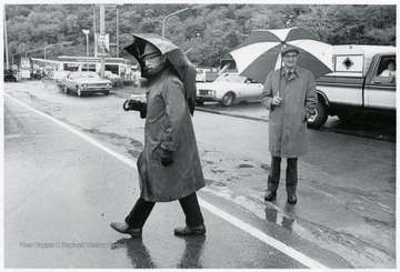 Two men with umbrellas stand on the road.