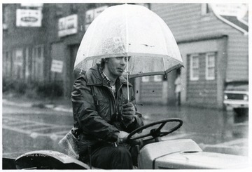 Man with an umbrella driving a lawnmower.