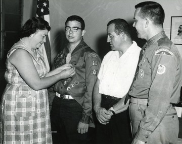 Mrs. Hartley (left) is pinning a medal (likely an Eagle Scout award) on her son.  Robert Hartley and Rev. LeJeune Lewis (far right) watch.