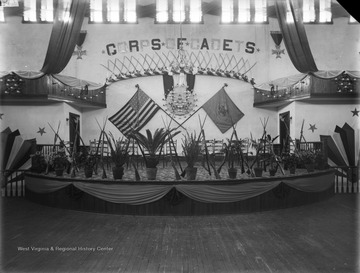 WVU Commencement Hall decorated with potted flowers and plants, garlands of lights, swags of fabrics and bows; on the backdrop the U.S. Flag and unidentified flag spread.  The Banner over the flags and a centerpiece of lights reads CORPS OF CADETS.