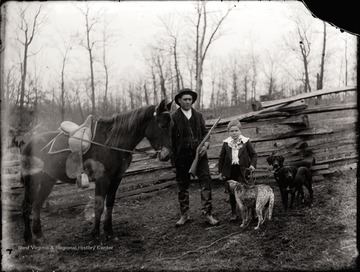 A portrait of man and boy; the father holds a horse rein and a gun; the son holds leashes for three dogs; the photo taken outdoor.