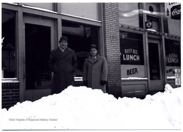 Two men stand on the sidewalk, Hewes Street behind piled snow.
