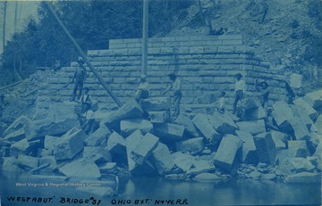 Unidentified workers stand among cut stones used in the construction of bridge abutments (such as one in the background). The poles in the photograph are part of the pulley used to place the stones.