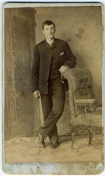 Dressed in the fashion of the late 1800s, posing with his arm resting on a chair.