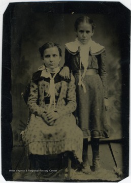Two young girls (probably 8 or 9 years old). They both have their hair pinned back and are wearing long dresses below the knee dresses in the fashion of the day for children.