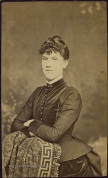 A young woman wearing a high collared top and long, bustled skirt. 