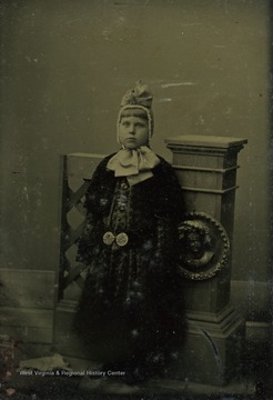 Unidentified young girl poses wearing a large outer garment, held at the waist with ornat belt, and a bonnet tied with a huge ribbon.