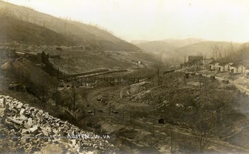 Railroad tracks run down the middle of the valley, while cleared mountainsides give way to industrial operations pictured on the left and the town buildings on the right.