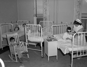 Nurse gives a drink to boy in hospital bed who is wearing a mask covering his eyes after apparent operation.