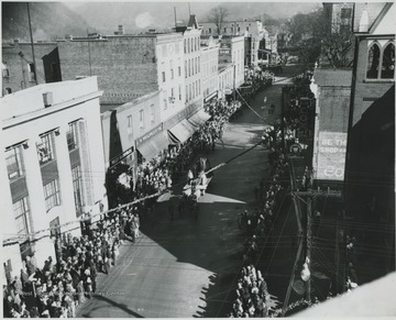 Crowds gather along the street to watch what appears to be a parade. Pictured in the background is First National Bank, H. H. Woolworth Co., G. C. Murphy Co., Willey Hardware, Westinghouse Appliances, and A. W. Cox Department Store. 