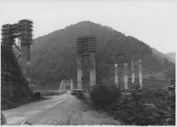 Large supporting structures tower over a smaller bridge connecting a dirt road. 
