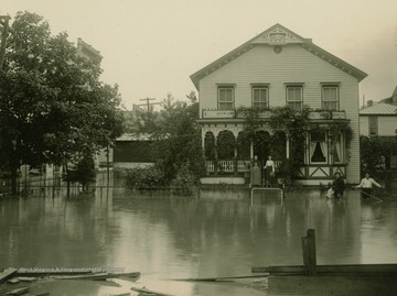 Two men stand in water up to their thighs with their dog standing on it's back feet along fence. Meanwhile the two women stay dry on front porch steps.