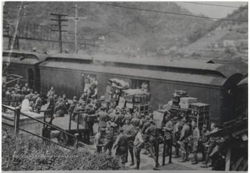 A crowd of men in uniform gather around the passenger train and load their luggage. 