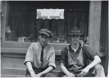 Two men, one in overalls, sit idly beside the building. 
