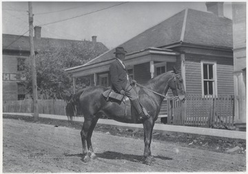 A man, who is probably Dr. Ryan, sits on top of a horse in the middle of a dirt road. The Hinton General Store can be seen in the background.