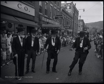 Four unidentified men wearing old-fashioned suits and full beards dance in the street while spectators watch from the sidewalks. 