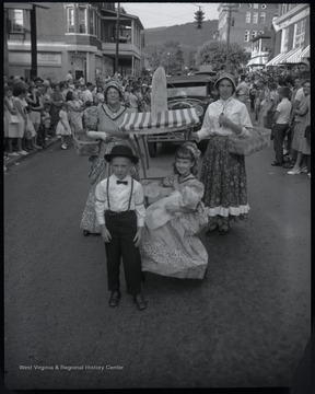 A young boy and girl pose in front of two older women in the middle of the street. They are dressed in old-fashioned attire to reflect the style of 1863. Spectators watch from the sidewalks. Subjects unidentified. 
