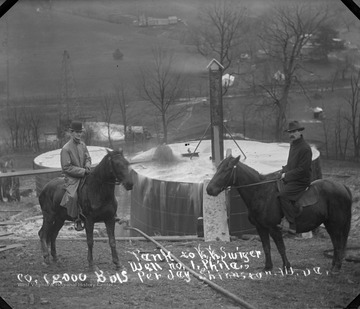 Two men on horses pose next to an oil tank.