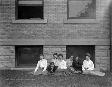 Two male and five female students of West Virginia University sit in the grass next to what is likely the Central Public School in Morgantown, W. Va.  The Central Public School was built in 1898-1899 and was opened in September 1899 to grade and high school students.