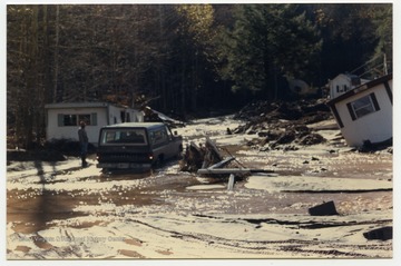 An unidentified man stands next to a car and several houses that were flooded when a stream, possibly Seneca Creek or White's Run, changed it's course during the 1985 flood in the area around Parsons, Elkins, Onego, and Mounth of Seneca, W. Va.
