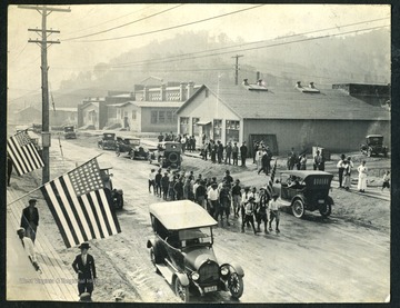 Unidentified boys and men march in a parade down the street in Nitro, with factories in the background. Nitro was created during WWI in 1917 to produce gunpowder for the war effort.