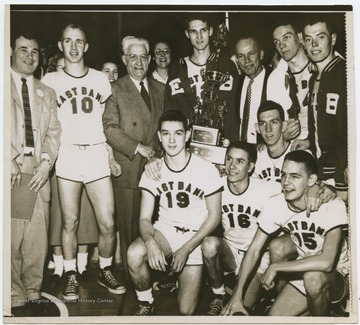 A portrait of the team, coached by Roy E. Williams. Jerry West is pictured in the center holding the trophy. The 1956 team secured the first ever state championship title for East Bank High School's basketball team.West was the team's starting small forward. He was named All-State from 1953–56, then All-American in 1956 when he was West Virginia Player of the Year, becoming the state's first high-school player to score more than 900 points in a season.