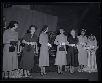 Fashionably dressed women stand beside each other in front of a stage. Subject unidentified. 