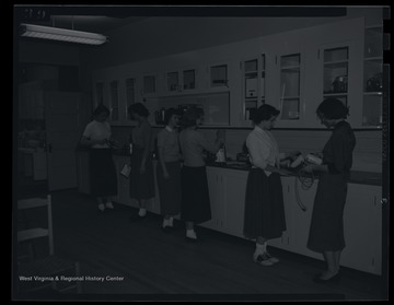 A group of girls are lined up along the counter and handling kitchen equipment. Subjects unidentified. 