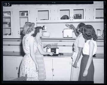 Four unidentified women look at a variety of electric kitchen appliances, including a crock pot and fryer. 