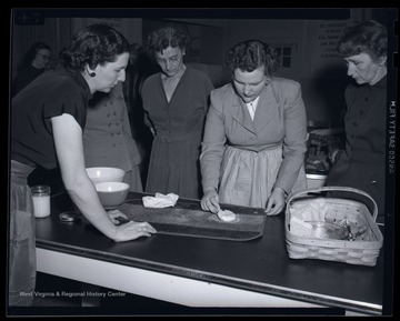 A group of women watch as a woman demonstrates how to remove a stain from a place mat. Subjects unidentified. 
