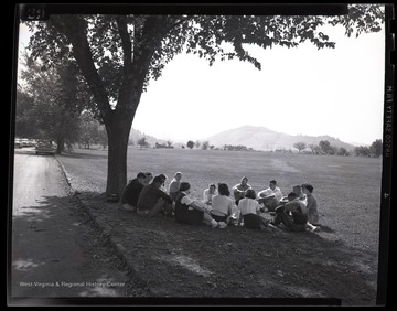 A group of boys and girls sit in the shade. Subjects unidentified. 