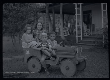 Miller Murrell, in a striped shirt, and three friends sit in a wooden jeep.  Behind them is a large porch with several adults sitting.  A large toy gun sits on the ground.