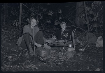 Miller Murrell and his mother cook with several pots and pans over a fire.  They appear to be camping outdoors.