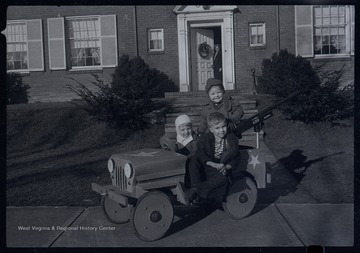 Miller Murrell and two other children sit in front of a house, likely on or near Ballengee Street in Hinton, W. Va.  The children are in a wooden jeep.  A large toy gun is mounted on the rear of the vehicle.