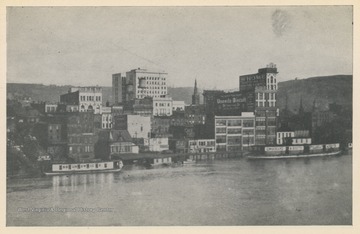 Photo postcard of the Wheeling waterfront during a flood.  Several buildings and business signs are visible, including the Schmulbach building, Uneeda Biscuit, The Home Outfitting Co., Chew Mail Pouch Tobacco, and others. Postcard is part of a souvenir book of 1913 flood images.