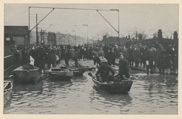 Photo postcard of a flooded street in Wheeling, W. Va. during a 1913 flood.  Several boats float in the flooded part of the street while crowds gather on dry sections. Postcard is part of a souvenir book of 1913 flood images.