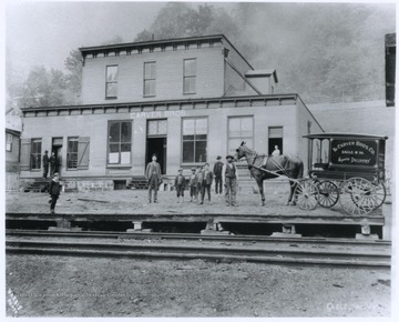 A horse-drawn carriage advertising the company is pictured on the right. A group of men and young boys are pictured in front of the store's entrance. 