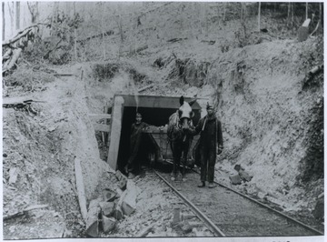 An African-American miner leads a horse and cart of coal out of a mine entrance.