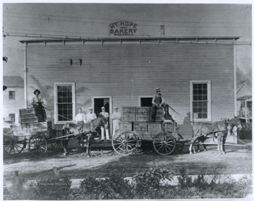 Horse-drawn carriages are loaded with crates outside of the bakery building, which advertises wholesale goods.
