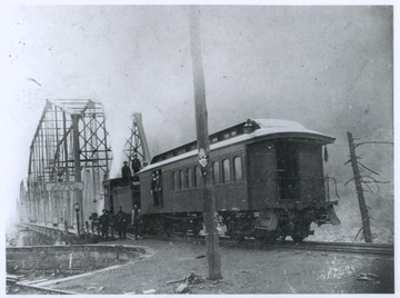 A group of men are pictured on and beside a train car.  A bridge in the background crosses the New River.