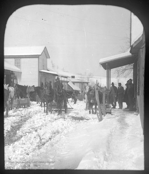View of the snow-covered street which is filled with horse-drawn carriages. A group of men stand beneath an awning on the right. 