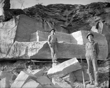 Two unidentified men pose beside the stone they are cutting large blocks out of. A long, chiseling tool rests against the rock. 