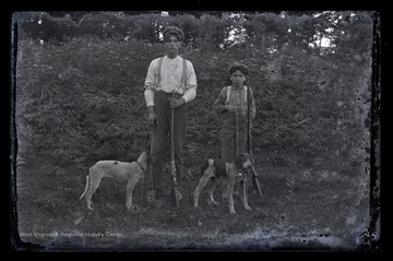 A young man and a boy pose with their hunting rifles. Beside them are two dogs. 