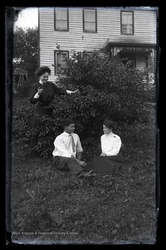 The woman dressed in black appears to be spying on the man and woman sitting beneath a bush.
